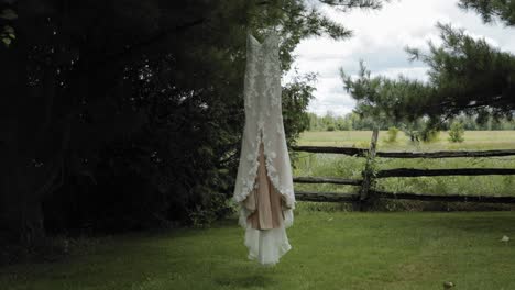 Elegant-designer-white-wedding-gown-hanging-from-the-pine-tree-in-the-back-yard-of-a-acreage-with-trees,-a-wood-fence-and-a-farm-field-in-the-background-at-the-Strathmere-Wedding-and-Event-Centre