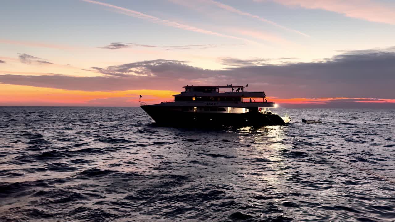 En majestætisk tredækket luksusdykkersafariyacht i silhuet mod en betagende orange solnedgang over det rolige Røde Hav en fredfyldt efterårsaften med gyldent time-lys og udsigt i øjenhøjde.