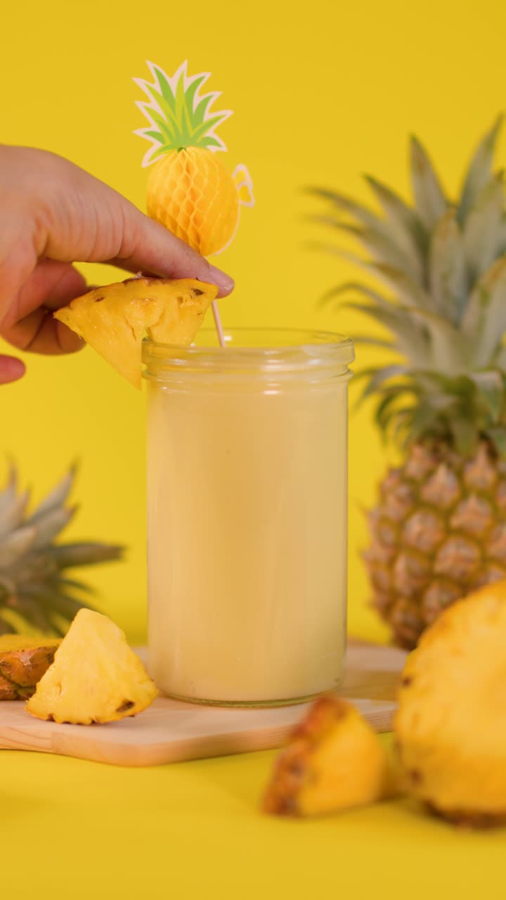 Una mano posiziona una decorazione a forma di ananas su uno smoothie cremoso, circondato da frutta fresca, su uno sfondo giallo brillante.