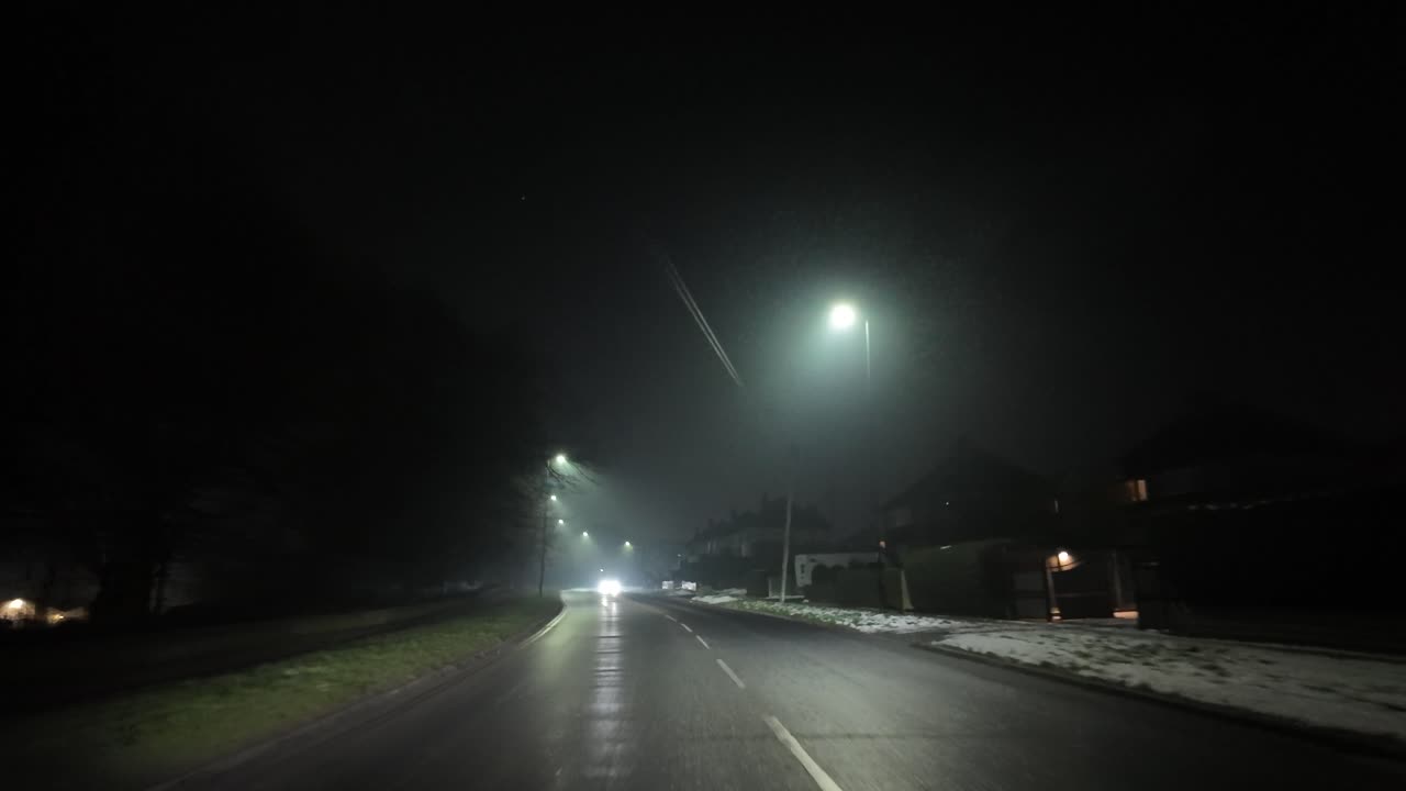 Dimly lit foggy road at night with glowing streetlights and snow along the sides.