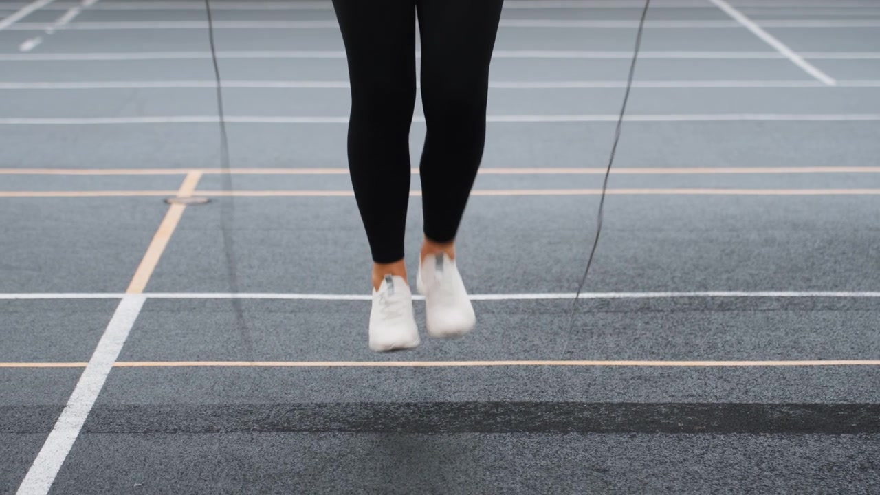Solo athlete practicing badminton footwork outdoors on lined asphalt court, white sneakers and black leggings visible, repetitive jump and landing drills, focused rhythm and balance, cool overcast
