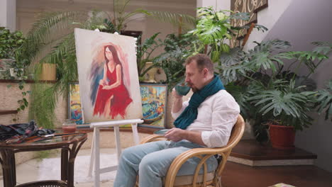 man gazes at portrait with cup of coffee in hand. artist appreciates juxtaposition of artwork and beverage creating moment of reflection in art studio