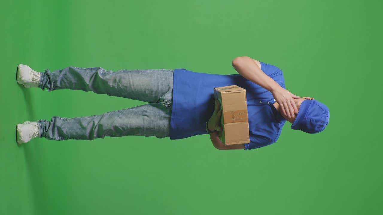 Full Body Of Tired Asian Male Courier In Blue Uniform Shaking His Head, Touching His Face, And Stretching While Delivering A Carton On Green Screen Background In The Studio