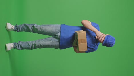 full body of tired asian male courier in blue uniform shaking his head, touching his face, and stretching while delivering a carton on green screen background in the studio