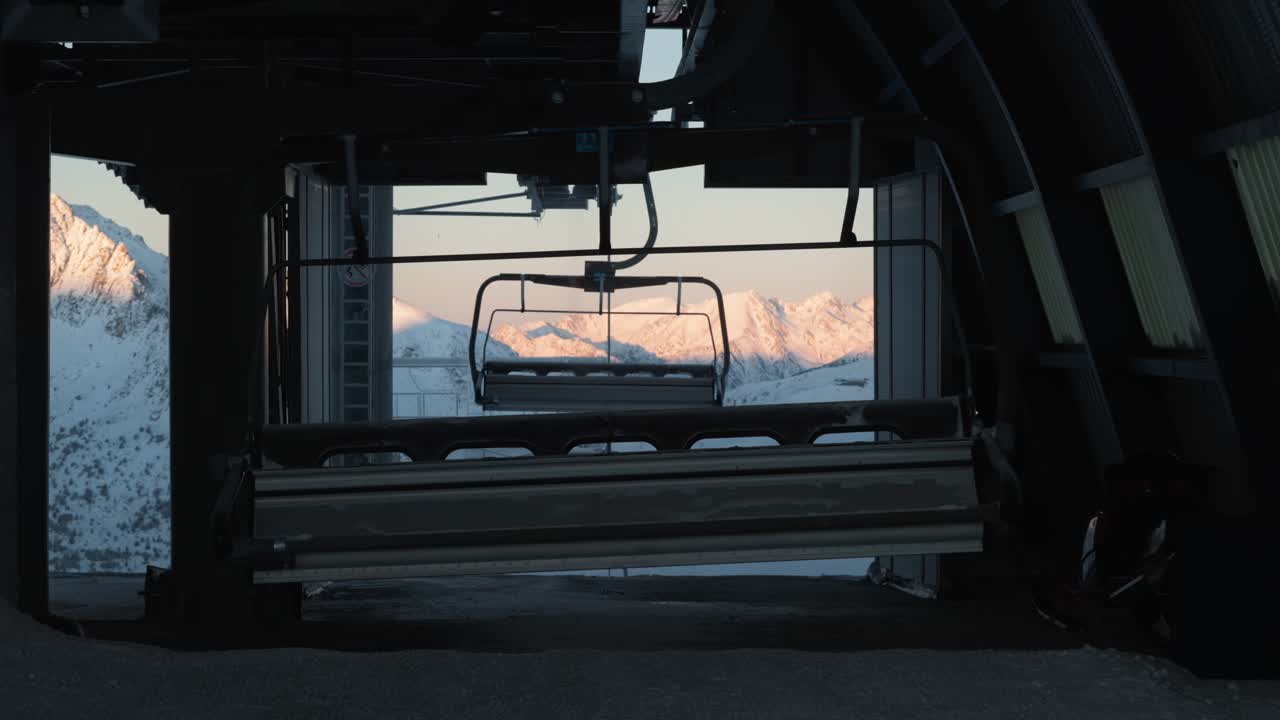 Chairlifts arrive and turn at the top station, shaded, with sunlit snowy mountains in the background in Andorra.