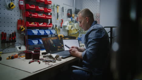 employee watching online trading lesson on laptop, checking real-time stocks, exchange market charts using smartphone and digital tablet. man remote learning trading during working hours in workshop.