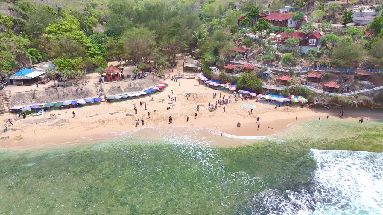 Kukup tourist beach on a sunny day, with people playing in the sand and the mountain with houses and dense vegetation in the background.