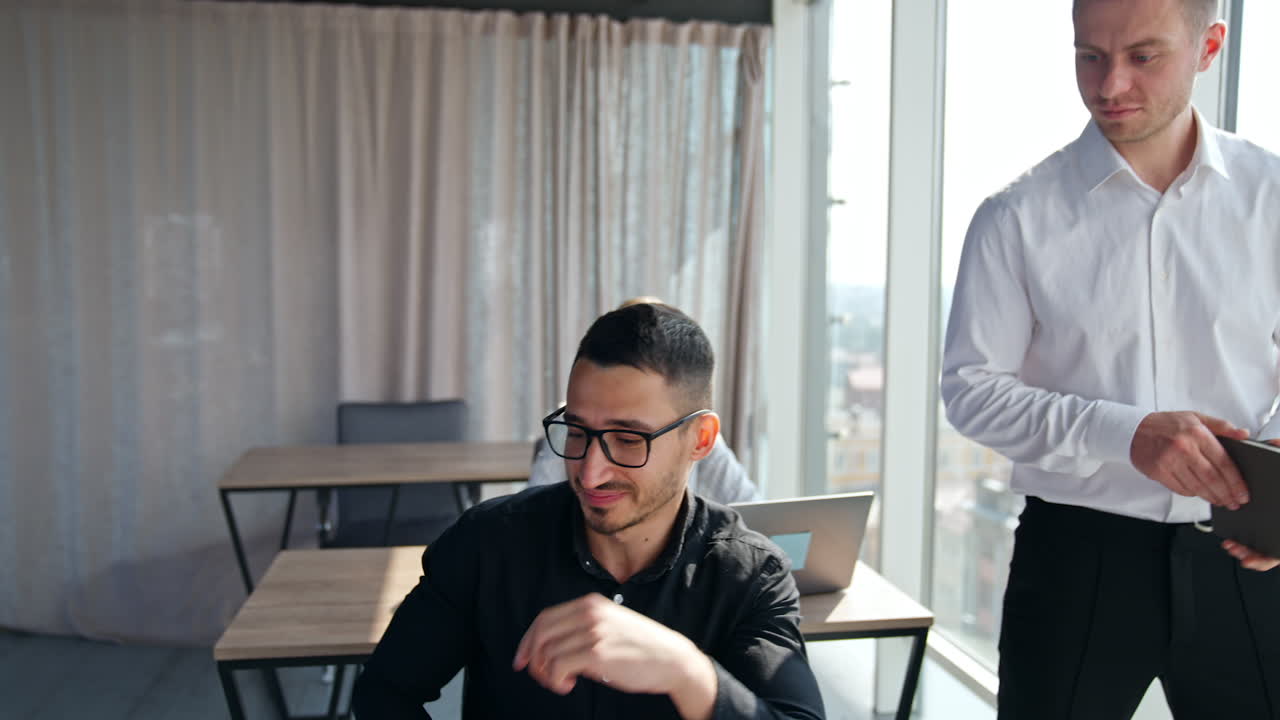 Man in glasses looks at the screen of his laptop. Manager in white shirt standing beside holding a paper notebook. Woman at backdrop works at computer.