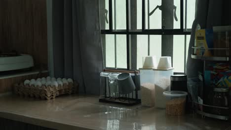 eggtray and toaster being placed on a cozy kitchen table by sunlit apartment window. -static wide shot