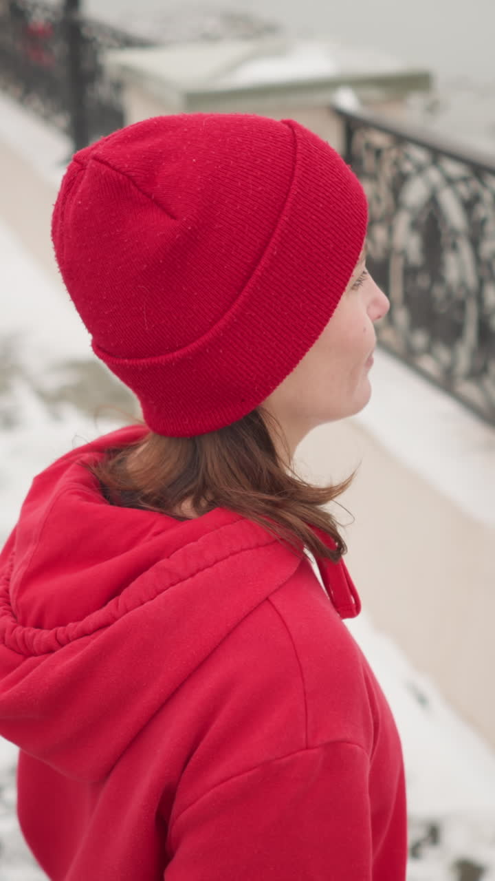 mujer con capucha roja y gorra camina hacia la valla de hierro descansando las manos en la barandilla decorativa, con vistas a aguas tranquilas rodeadas de tierra cubierta de nieve, reflejando una atmósfera de invierno pacífica y serena