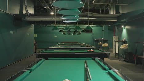 shot of green pool tables with billiard balls arranged on them and cue sticks placed across the tables. overhead lamps provide soft lighting, creating a cozy atmosphere in billiard room