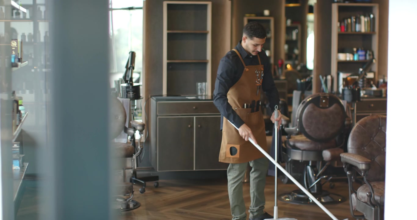 Afroamerikansk barber i forklæde ser åbent-skilt og mopper butiksgulv med moppe, forbereder butikken