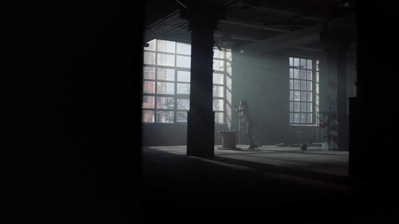 En pige udfører et sæt box jumps i et træningslokale af loftstypen. En kvinde træner i fitnesscenteret.
