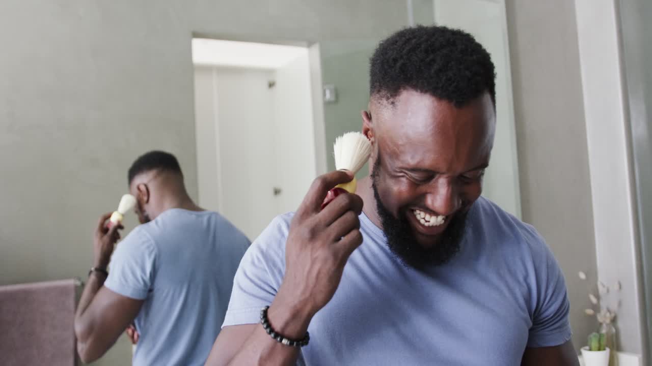 Afro-Amerikaanse man pakt gezichtsborstel, gebruikt deze op zijn wang bij de kaptafel, tikt op zijn telefoon en leest berichten.