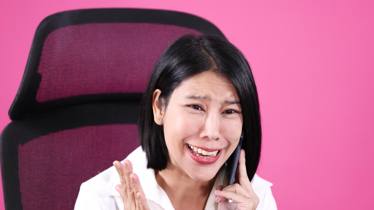 Young woman in office chair expresses shock and joy on phone, bright lighting, pink background