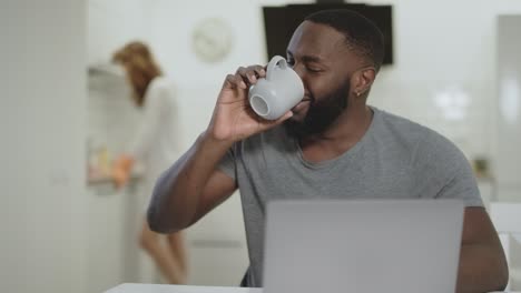 Hombre-Negro-Riendo-Bebiendo-Té-En-La-Cocina-Abierta.-Mesa-De-Limpieza-De-Mujer-Blanca