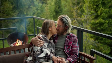 the blonde girl comes up to her boyfriend and sits down on the couch beside him. couple hugging and posing against the backdrop of mountains and forest