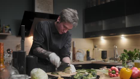 young professional male chef in an elegant black shirt with an alternative look, tattoos chopping fresh zucchini a modern, fancy looking kitchen with fresh vegetables besides cinematic sliding slo mo