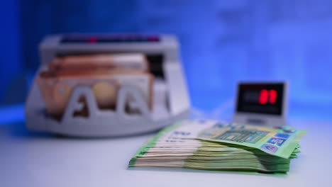 a stack of green banknotes lies on the desk. money counting machine counts fifty euro bills at blurred backdrop and put by the female hand at the foreground. close up.