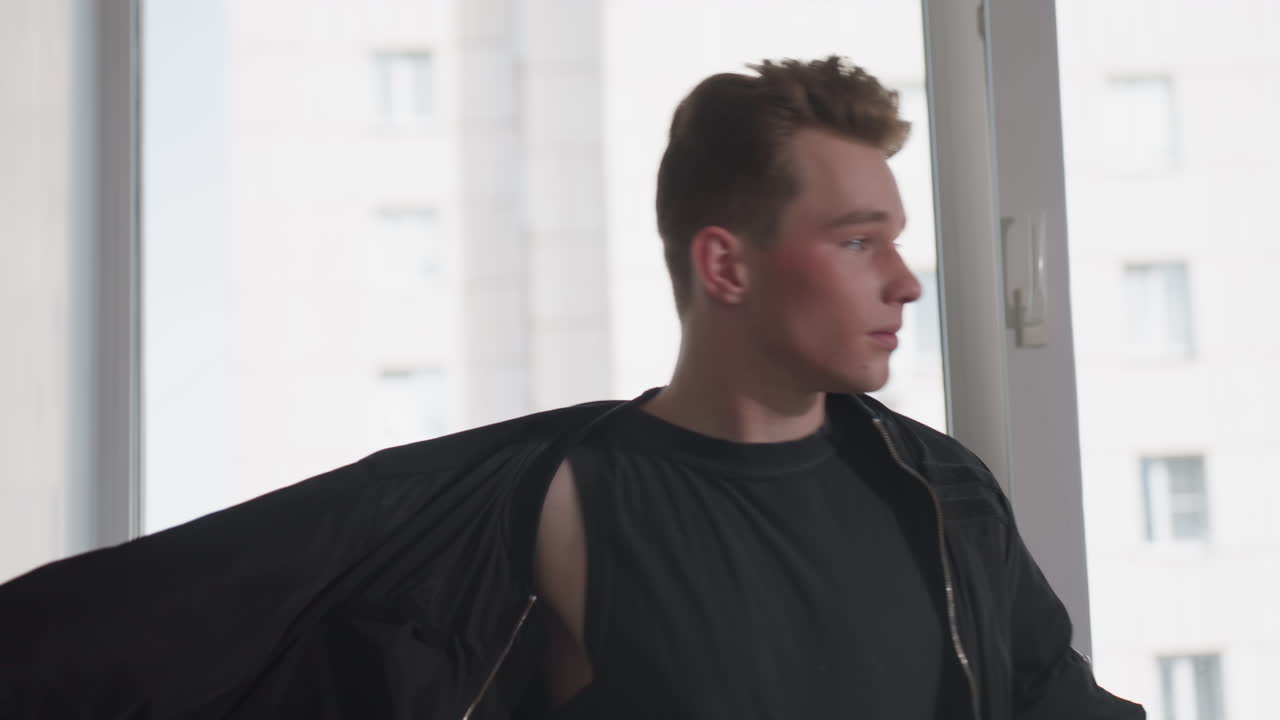 Young man in black tank top and high-waist trousers slips arms into black jacket sleeves while standing by large window overlooking city high rise, adjusting fit before heading out