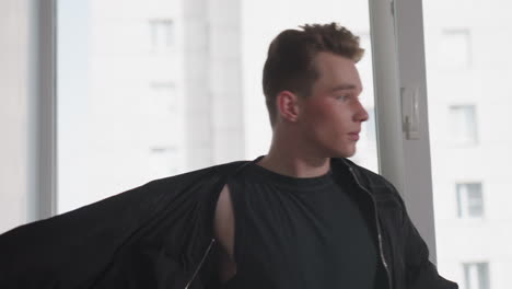 young man in black tank top and high-waist trousers slips arms into black jacket sleeves while standing by large window overlooking city high rise, adjusting fit before heading out