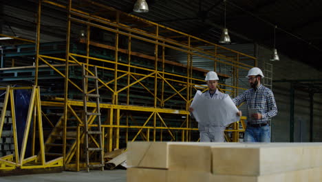 two men wearing helmets talking and walking in a factory with blueprint and tablet in their hands