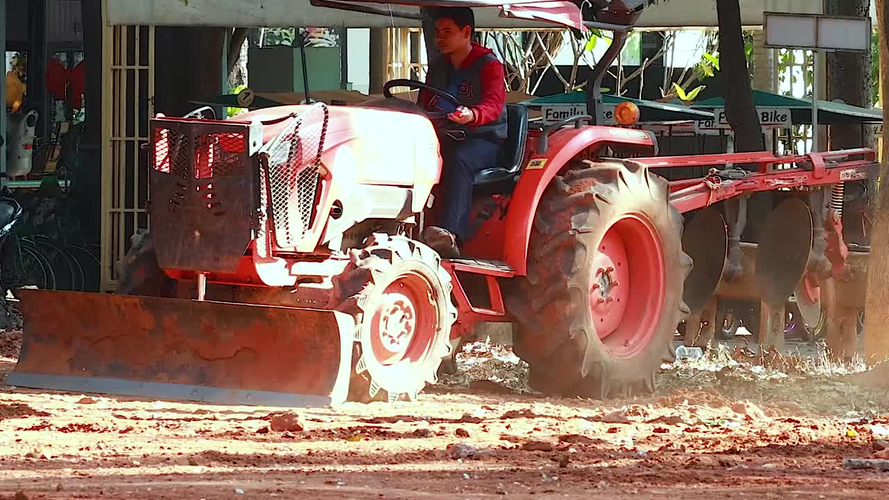 A red tractor operates on dusty soil, leveling the ground in a sunlit outdoor setting.