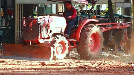 a red tractor operates on dusty soil, leveling the ground in a sunlit outdoor setting.