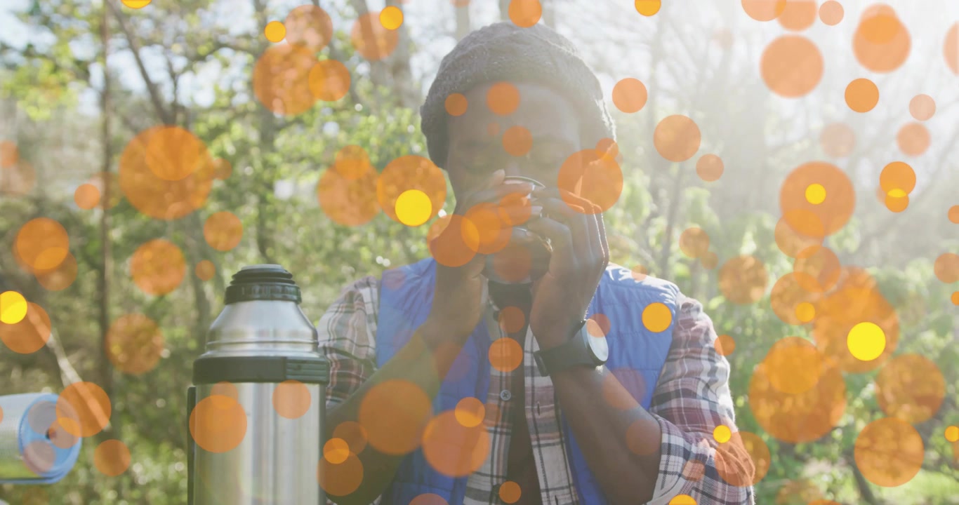 Uomo che svita il tappo del thermos e sorseggia una bevanda calda da campeggio, assaporando il calore mentre un bokeh arancione fluttua