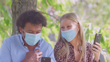 couple wearing masks meeting in outdoor park during health pandemic looking at mobile phones