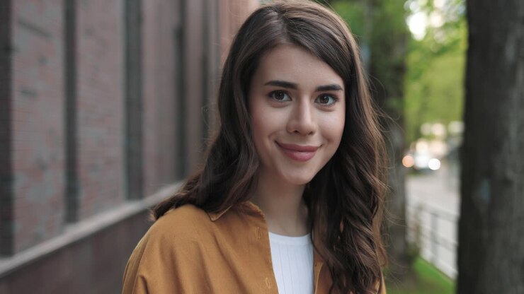 Portrait view of caucasian female student sitting in the street and smiling to the camera while relaxing during a break near the college