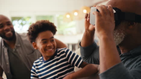 Abuelo-Usando-Auriculares-Vr-Mientras-Una-Familia-Masculina-De-Varias-Generaciones-Se-Sienta-En-El-Sofá-En-Casa-Juntos