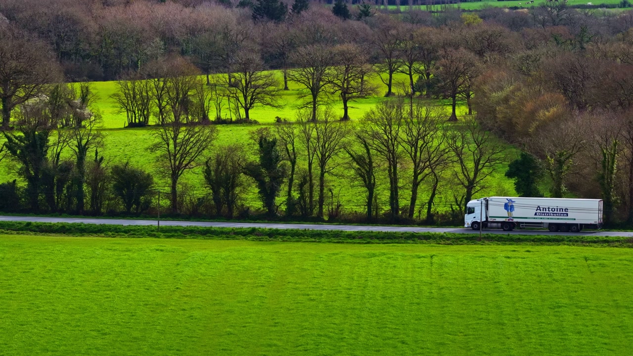 A truck driving along a rural road through bright green fields and tree-lined farmland, capturing countryside transport, spring landscape, and peaceful agricultural scenery.