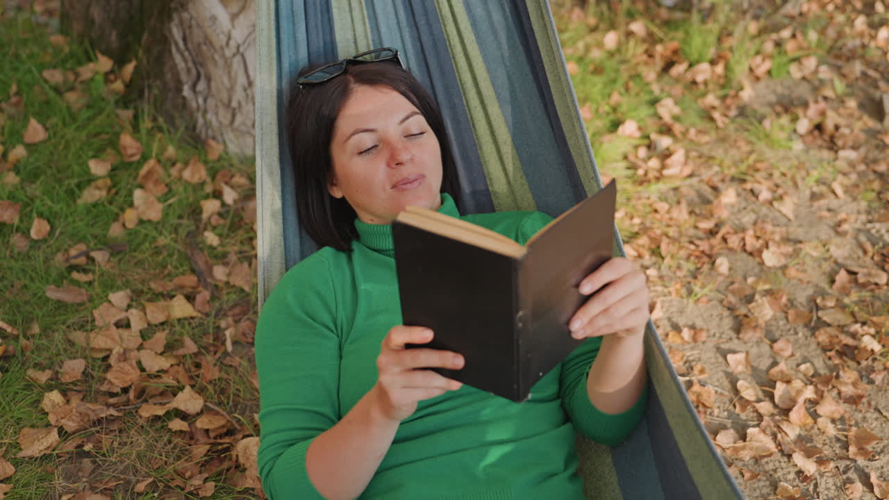 Witte vrouw die aan hangmatkamperen doet en hardop leest, met expressieve gebaren en dramatische vertelling, groene trui, gevallen bladeren onder de voeten, zonnebril opgezet, stem draagt door het bos, met artiestenenergie.