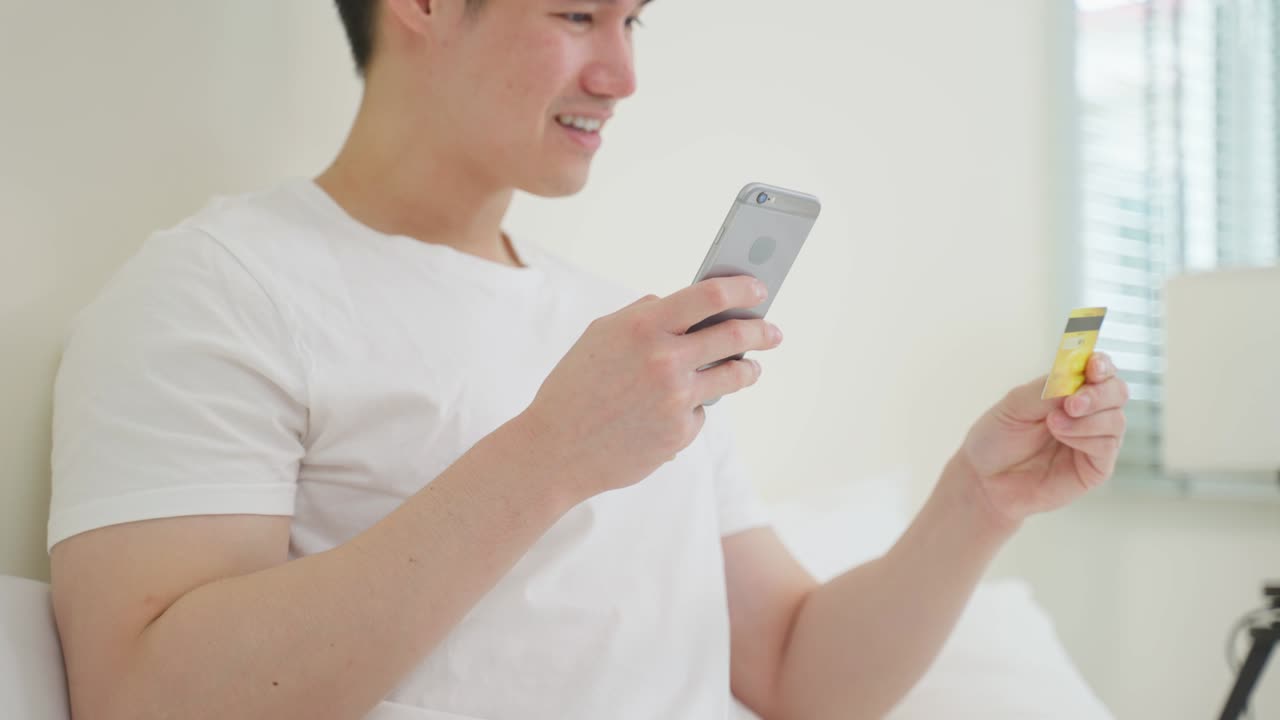아시안 매력적인 남성은 아침에 집에서 침대에서 온라인 쇼핑을 즐긴다. 예쁜 남자는 침실에 누워있고, 신용카드를 들고 은행에서 온라인 지불을 할 수 있습니다.