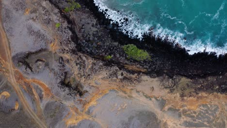 volcanic rocks and sandy terrain meet bright turquoise waves along the rugged coast. aerial view highlights erosion patterns and dramatic textures.