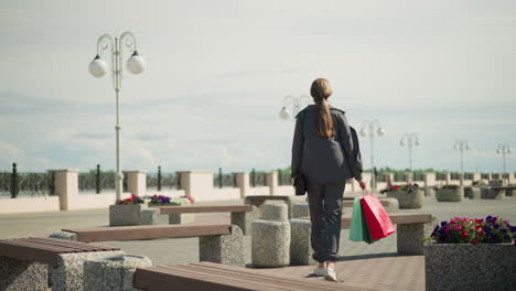 white lady walks up to outdoor bench carrying shopping bags in hand, she drops the bags onto the bench, and the mint-colored bag falls down, nearby, flowers and benches line the public space