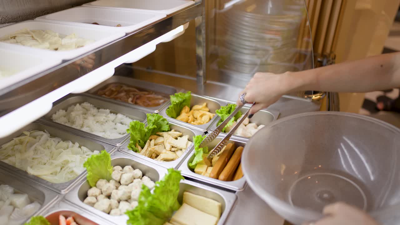Person uses tongs to pick raw hotpot ingredients from buffet under bright indoor lighting