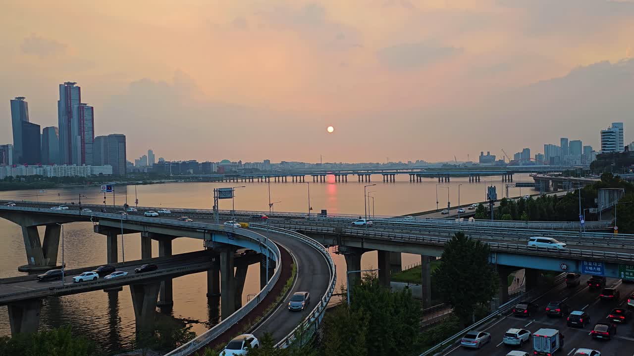 Busy city traffic as cars merge and exit on Wonhyo Bridge over the Han River at sunset with orange soft cloudy sky, Seoul's skyline in background, zoom out
