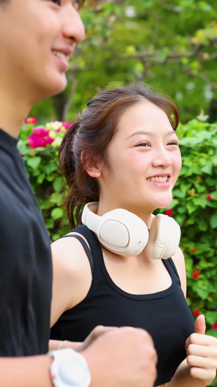 晴れた公園をジョギングする、スポーツウェアとワイヤレスヘッドホンを着用した幸せそうなアジア人カップル。