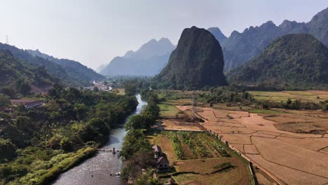 aerial view of pha tang in vang vieng, laos, revealing a lush river valley bordered by golden fields, rugged limestone mountains, and layered forested hills under a clear blue sky