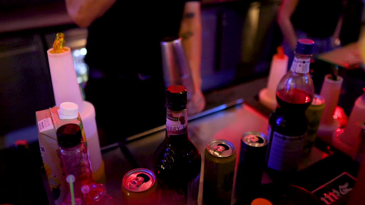 Bartender scoops ice behind bar counter, colorful bottles and cans, low light, nightclub atmosphere
