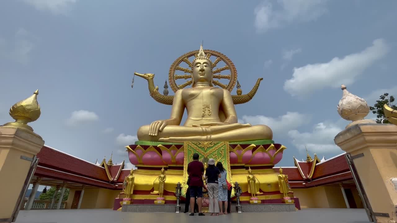 Captivating drone shot of a grand golden Buddha statue with intricate halo and symbolic gestures, ascending stairs, bright sun and blue sky in Ko Samui, Thailand