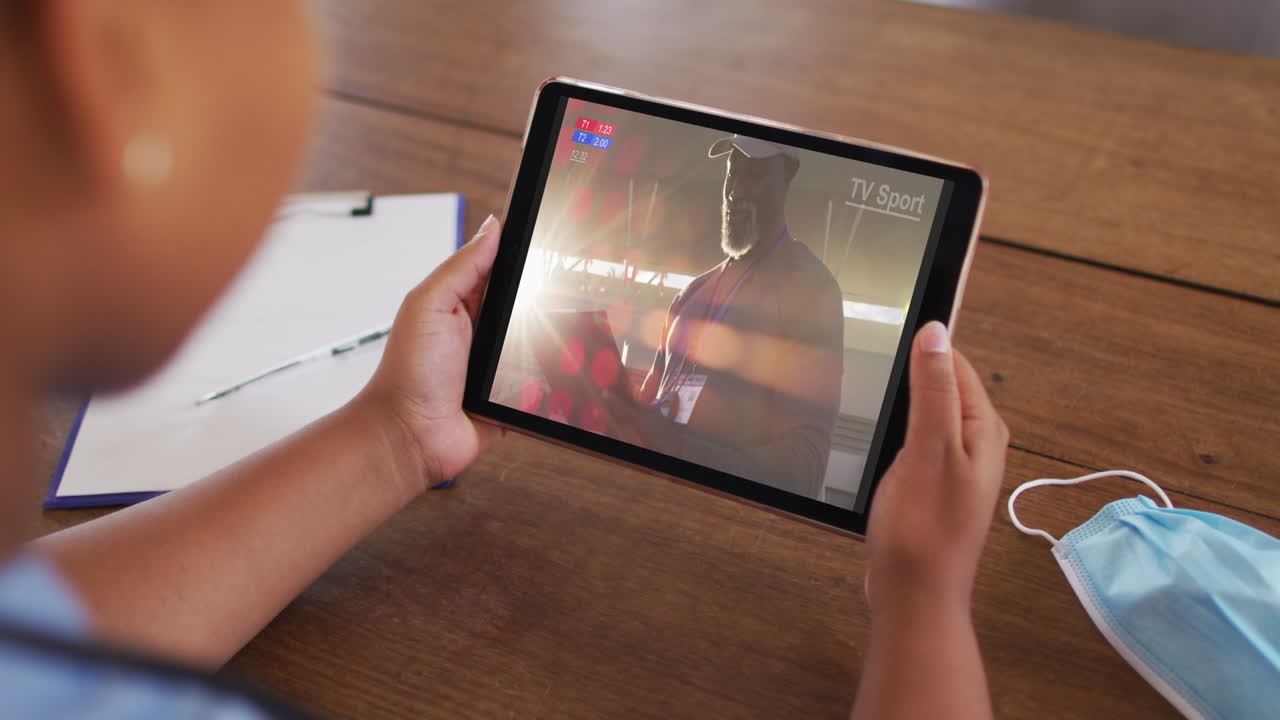compuesto de una mujer sentada en una mesa con una máscara facial, viendo un evento deportivo en la tableta