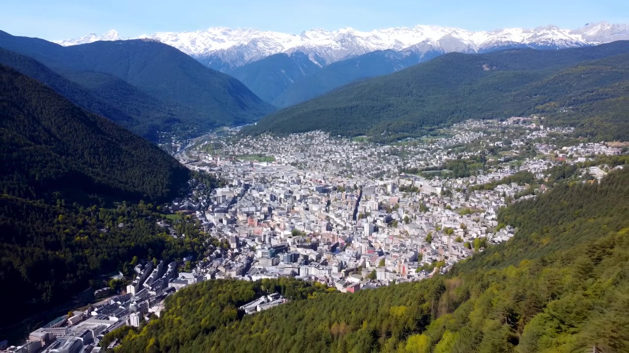 Weite Landschaftsansicht bewegt sich sanft über kompakte Stadtviertel, umgeben von bewaldeten Berghängen, wobei geschichtete Bergkämme und helle schneebedeckte Gipfel bei klarem Tageslicht sichtbar werden.