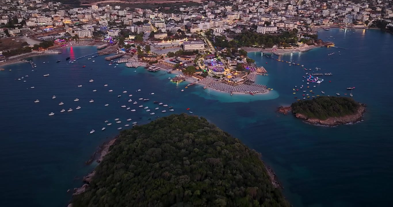 Vista aérea das ilhas de Ksamil e cidade costeira ao crepúsculo, Albânia. Imagem panorâmica de drone de pequenos ilhéus verdes, baía marítima com barcos e resorts de praia iluminados ao anoitecer