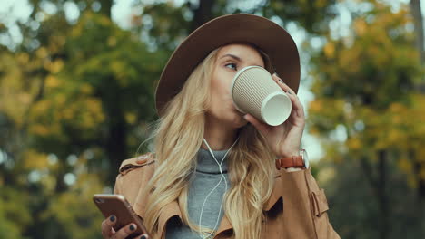 Caucasian-young-blonde-woman-in-a-hat-and-headphones-listening-the-music-on-the-smartphone-and-drinking-coffee-in-a-beautiful-autumn-park