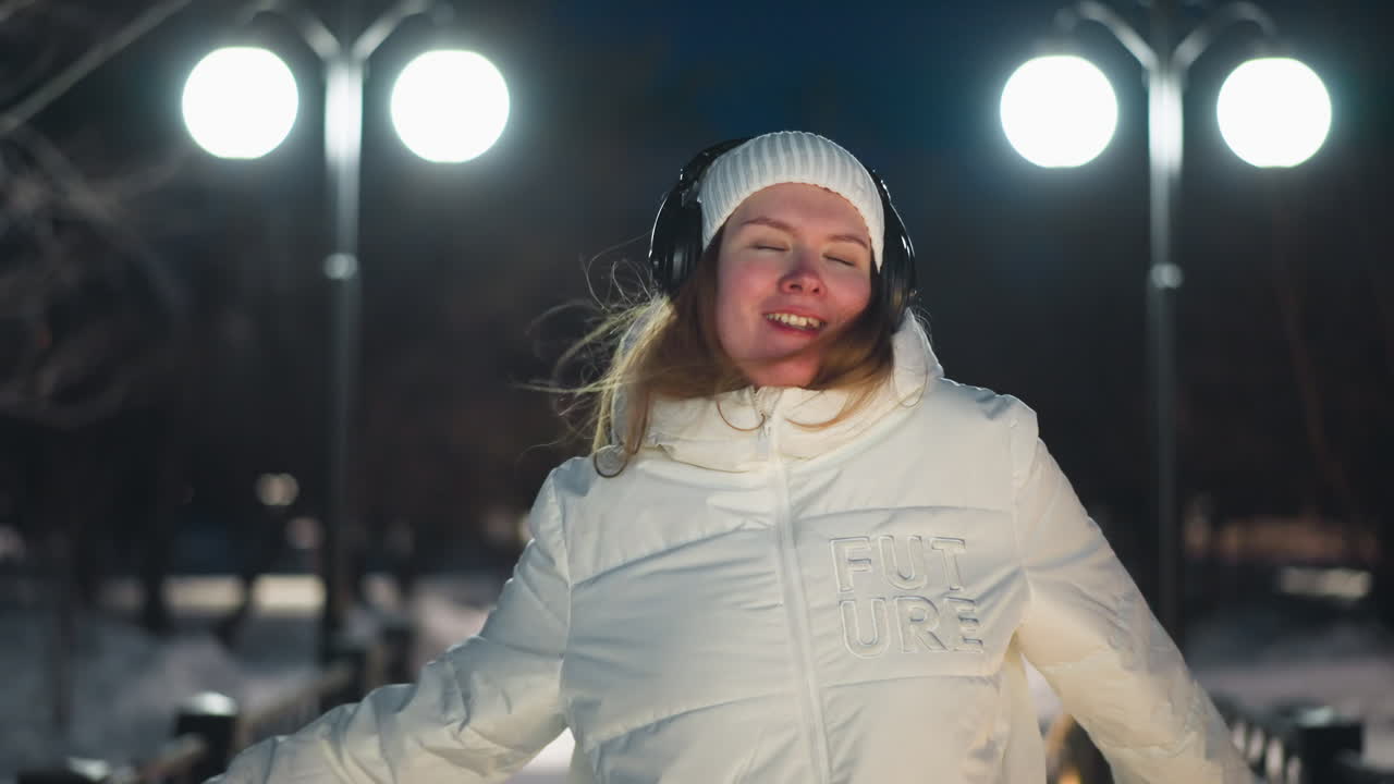 Lady with headphones and white puffer coat smiles and moves her body under bright streetlights on snowy pathway at night, joyful expression and dynamic rhythm in serene winter park