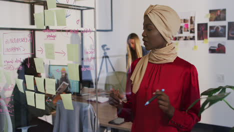 Mujer-De-Negocios-Afroamericana-Hablando-Con-Colegas-Y-Moviendo-Notas-Sobre-Una-Pared-De-Vidrio-En-La-Oficina