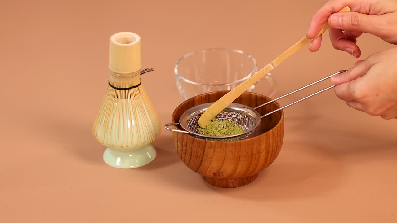 Hand sifts matcha powder through fine mesh into wooden bowl, minimal Japanese tea preparation setup